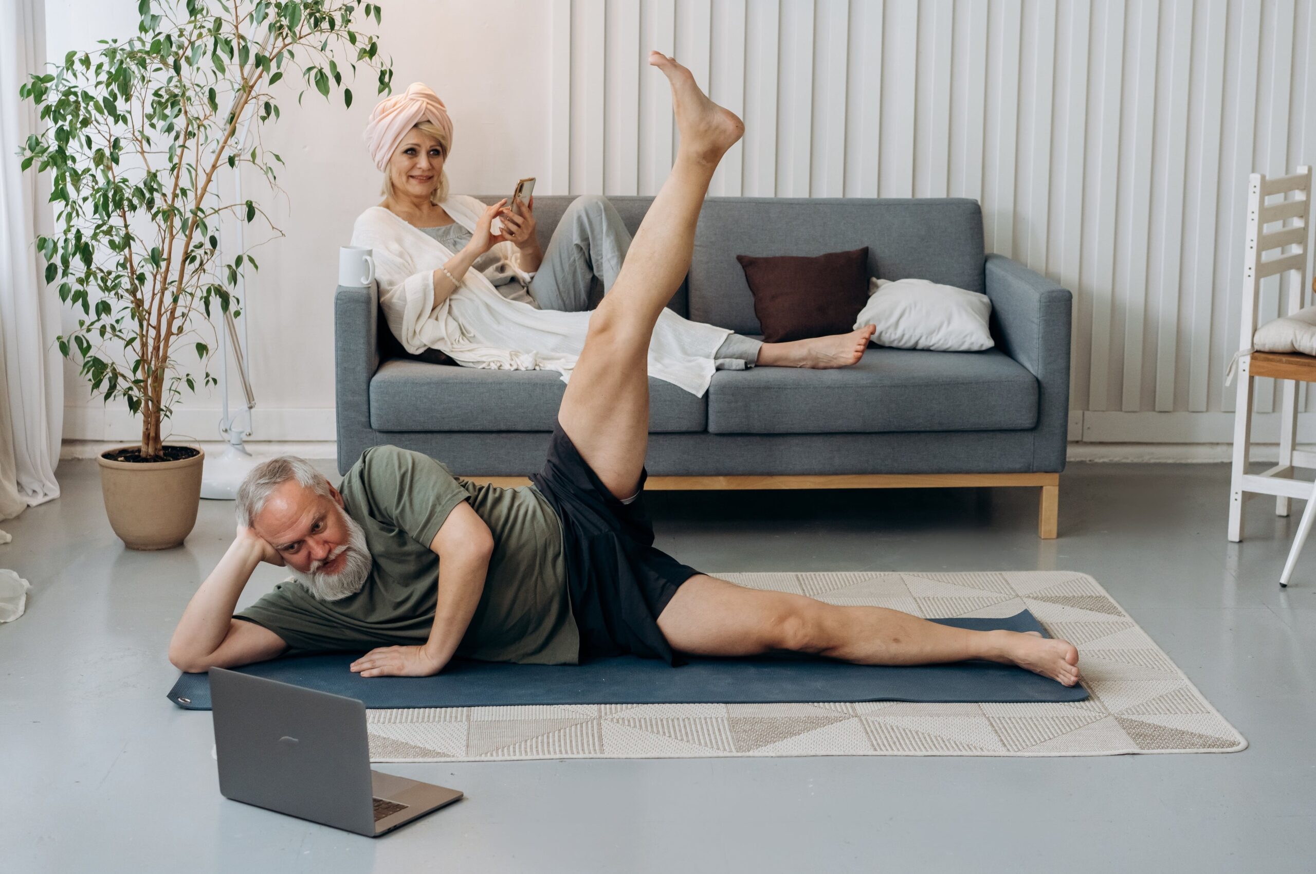 Older couple with woman sitting on sofa texting and man exercising while watching a laptop.