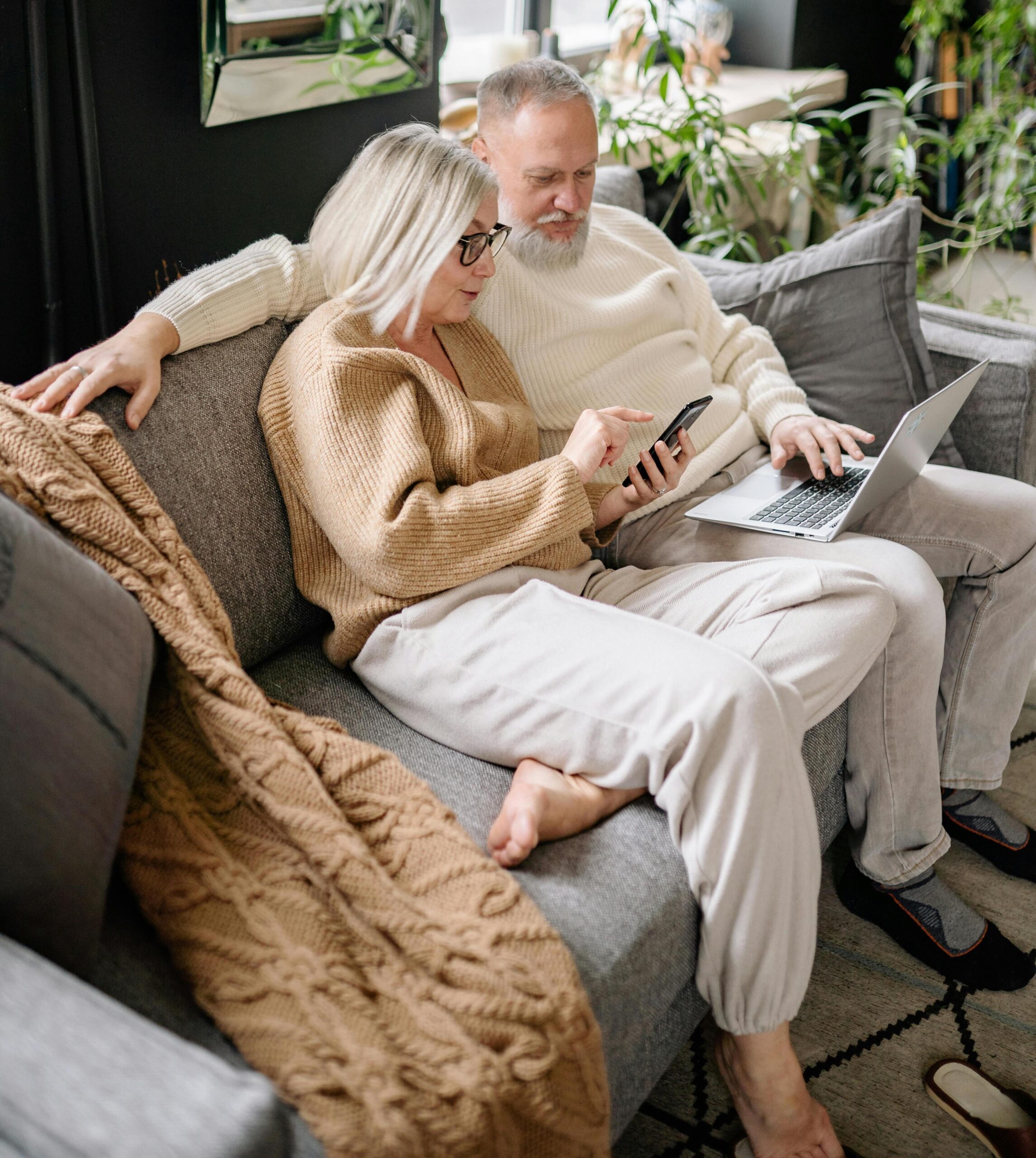 Older couple seated side by side on a sofa. The woman is using a smartphone and the man a laptop.